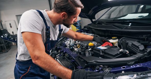 Man auto technician looking carefully inside car engine and touching its battery with hand during examination